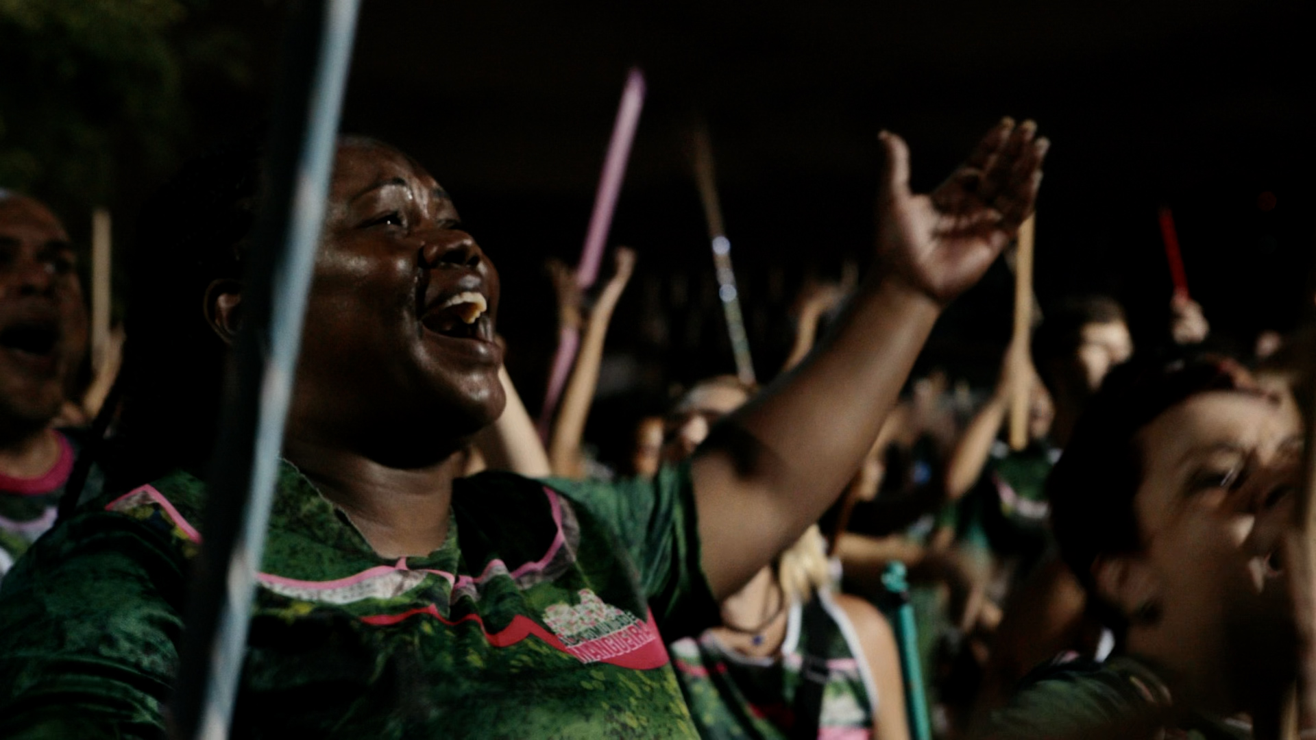 Foto que muestra a un grupo de personas en actitud de protesta. En primer plano aparece una mujer negra que parece estar entonando un canto o una arenga. Alza sus brazos abiertos y lleva en una mano un palo.
