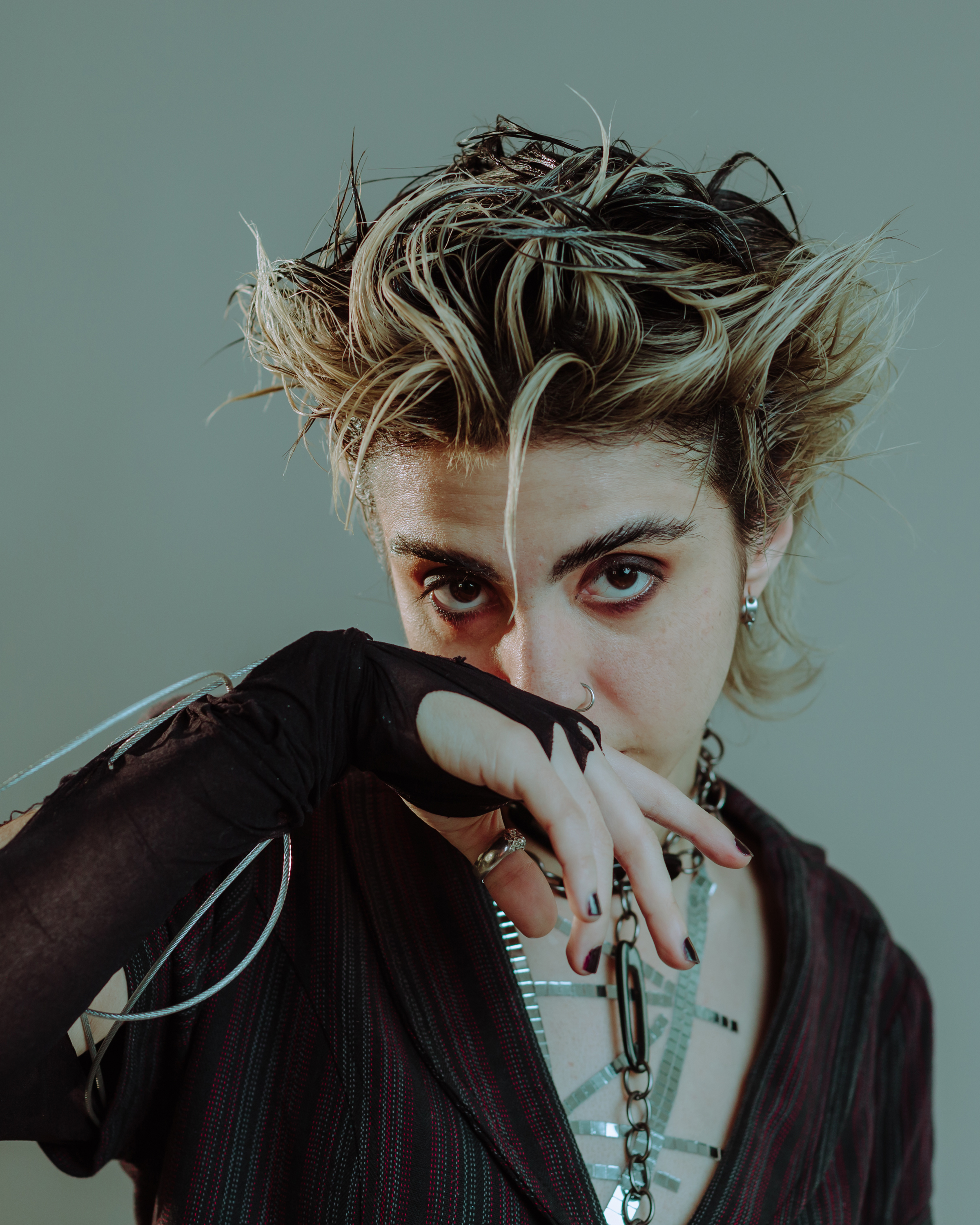 Foto de una chica joven con el pelo corto rubio, grandes ojos maquillados de negro y ropa y accesorios de estilo rockero, que mira de frente a la cámara con un brazo enguantada y la mano posada delante de su boca. También lleva las uñas pintadas de negro.