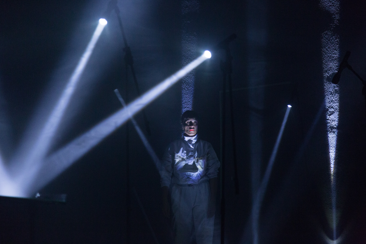 Foto de un espacio lleno de sombras y aces de luces que salen de varios focos. En medio de ese claroscuro hay una mujer parada en la que resaltan ...