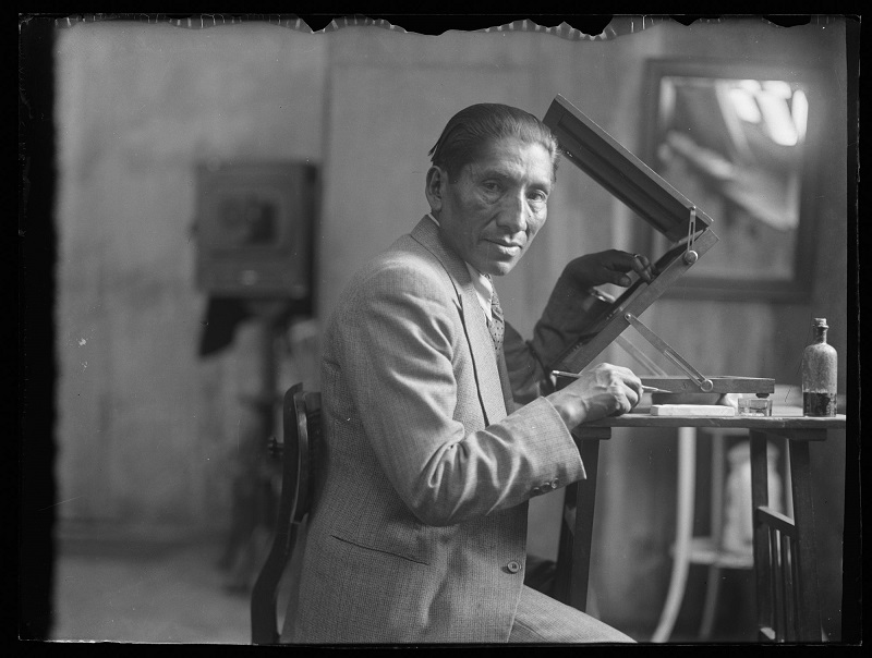 Fotografía en blanco y negro del fotógrafo Martín Chambi en su estudio, sentado ante una mesa de trabajo donde tiene un aparato de visualización ...