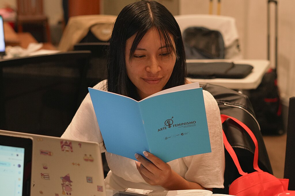 Foto de una joven snetada ante una laptop y leyendo un libro que en su portada dice Arte + Feminismo.