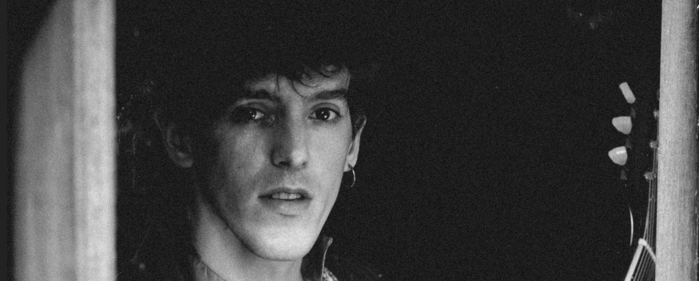 Foto en primer plano, en blanco y negro, de un joven que mira a la cámara con gesto melancólico y aspecto rockero. Lleva una melena ondulada, una argolla en una oreja y una guitarra eléctrica.