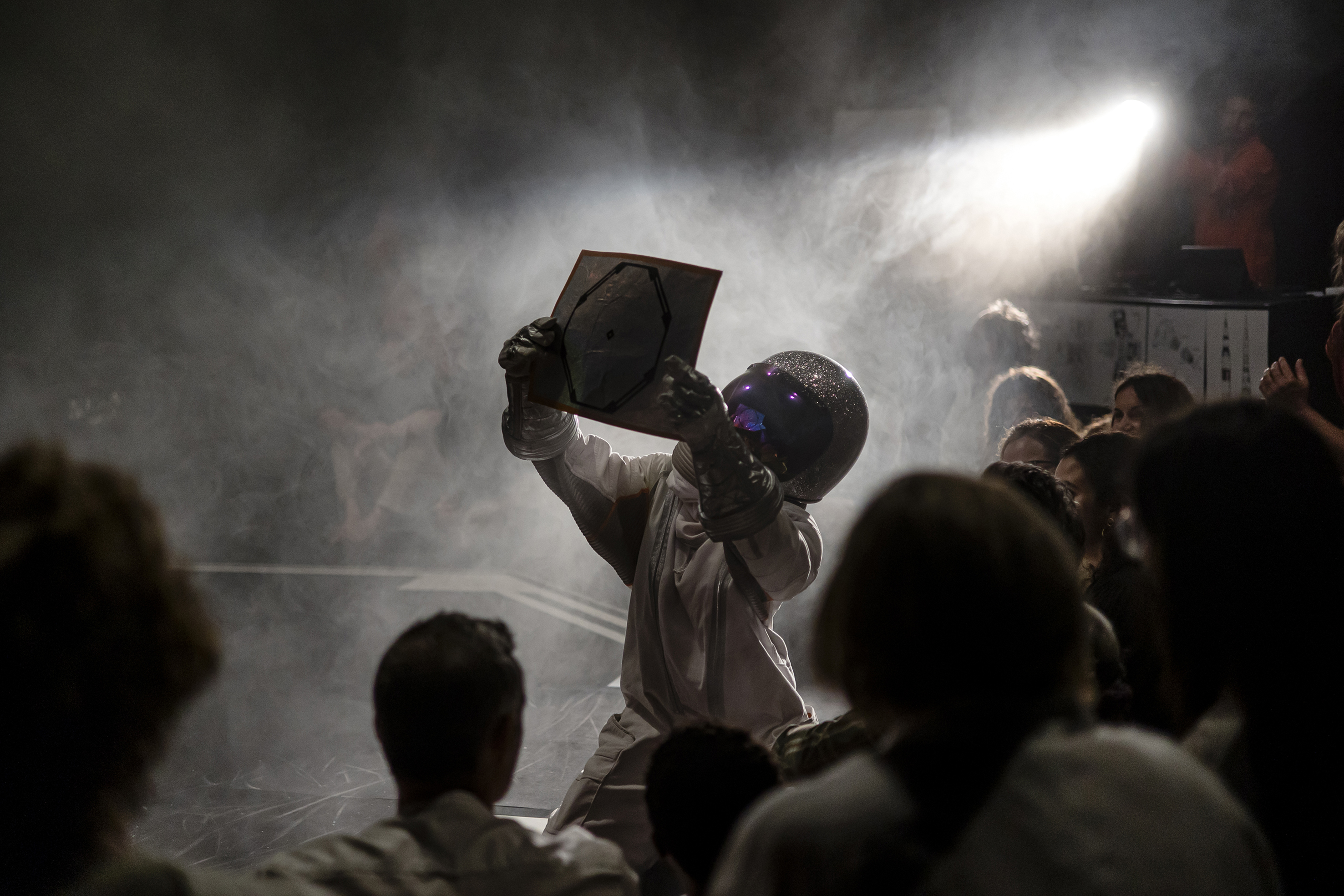 La imagen muestra a una persona vestida con escafandra y casco de cosmonauta, arrodillada en medio de un escenario circular y lleno de humo, mientras con las manos en alto una lámina del planeta tierra que le muestra a un público mayoritariamente infantil.