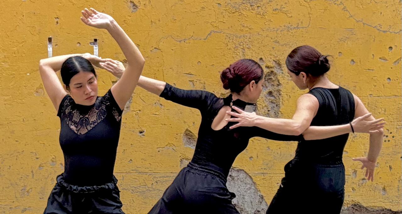 Foto que muestra, delante de una pared amarilla, tres bailarinas vestidas de negro con sus cuerpos entrelazados.