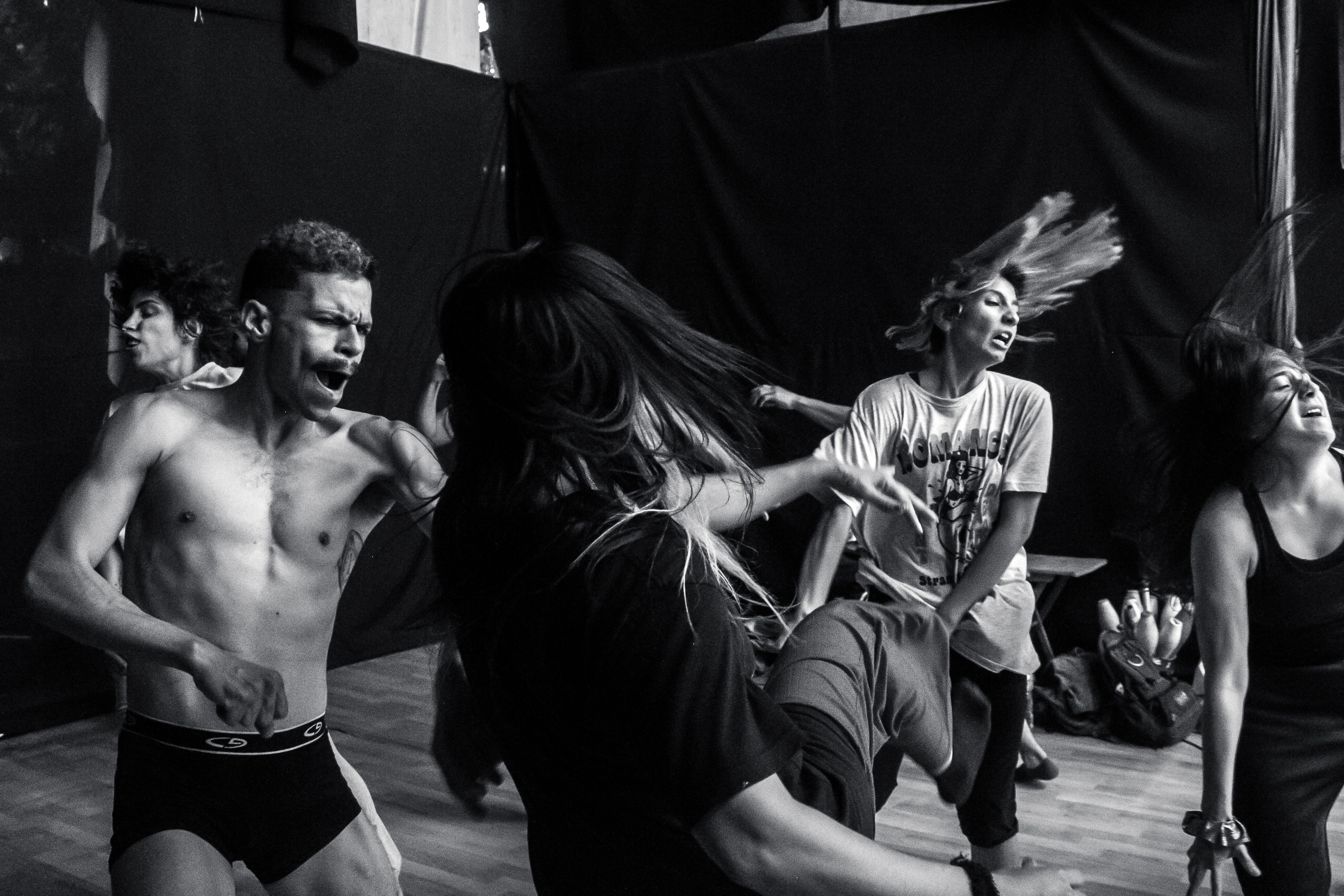 Foto en la que se ven a diferentes bailarines y bailarinas realizando enérgicos ejercicios de danza que hacen volar sus cabellaos. Se encuentran en un espacio rodeado de paredes de telas negras.