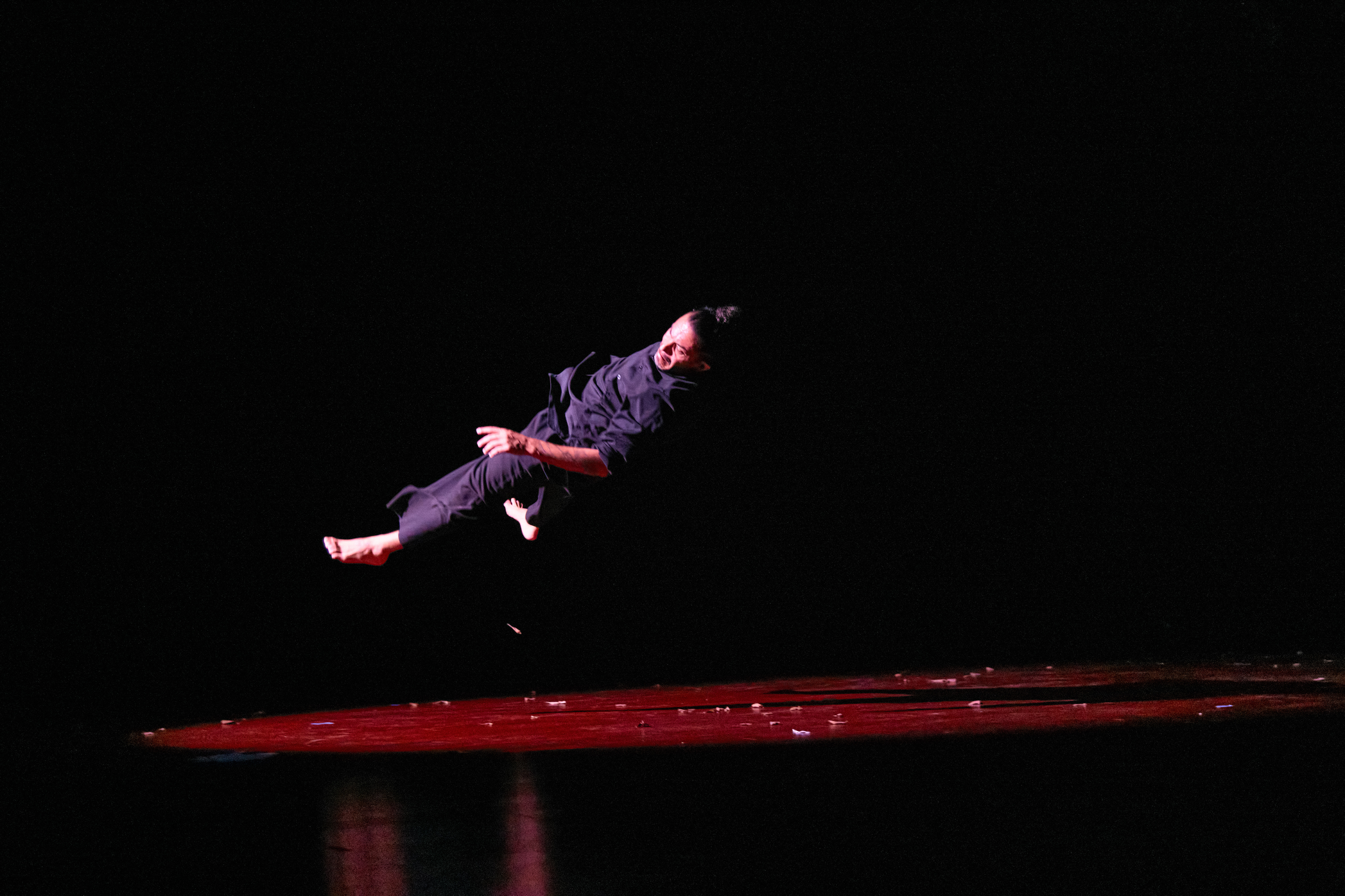 Foto que muestra a un bailarín todo vestido de negro dando un salto que lo sitúa en una posición casi paralela al suelo.