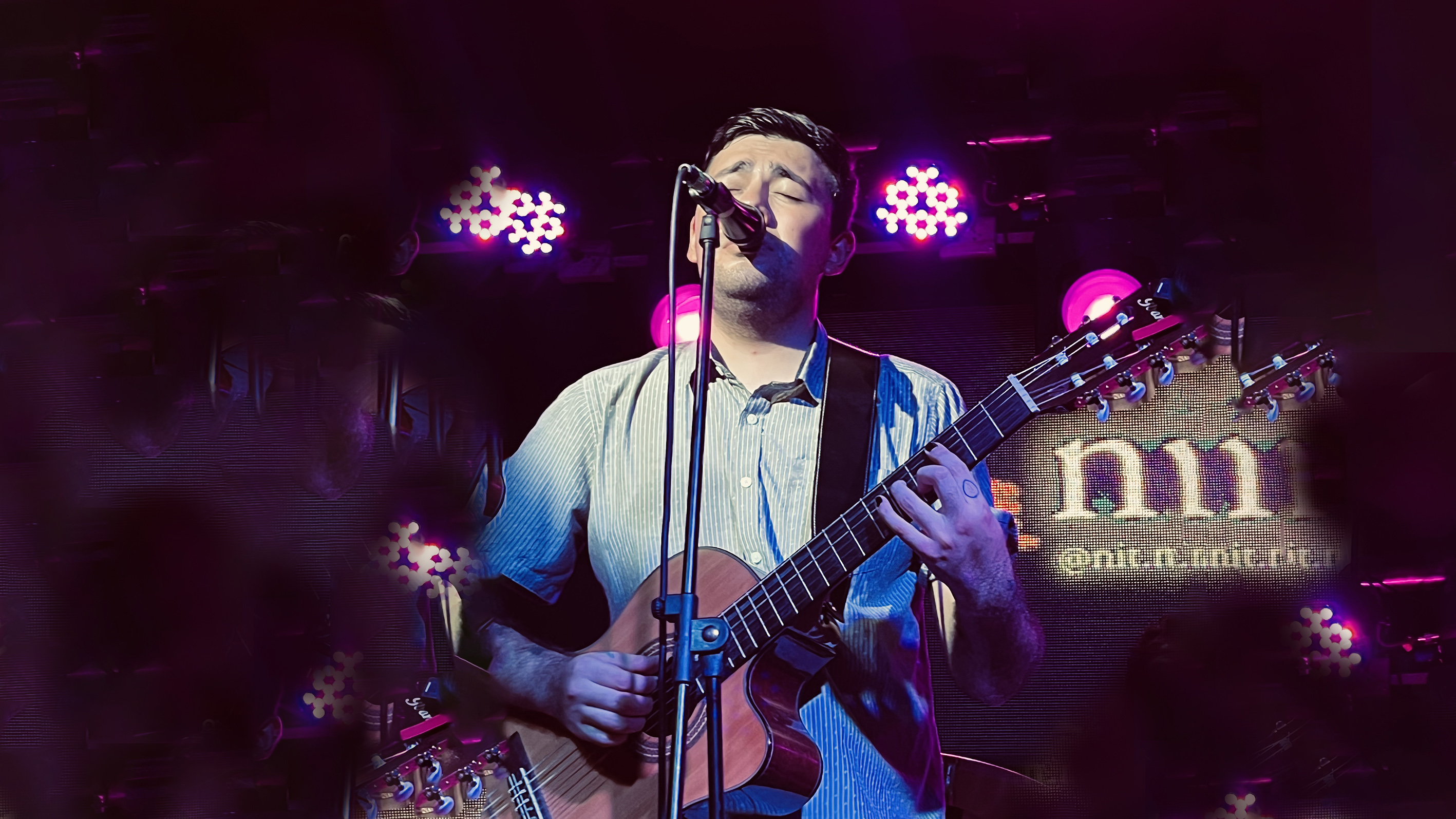 Foto de un joven cantando y tocando guitarra ante un micrófono de pie en un escenario rodeado de luces brillantes tipo discoteca.