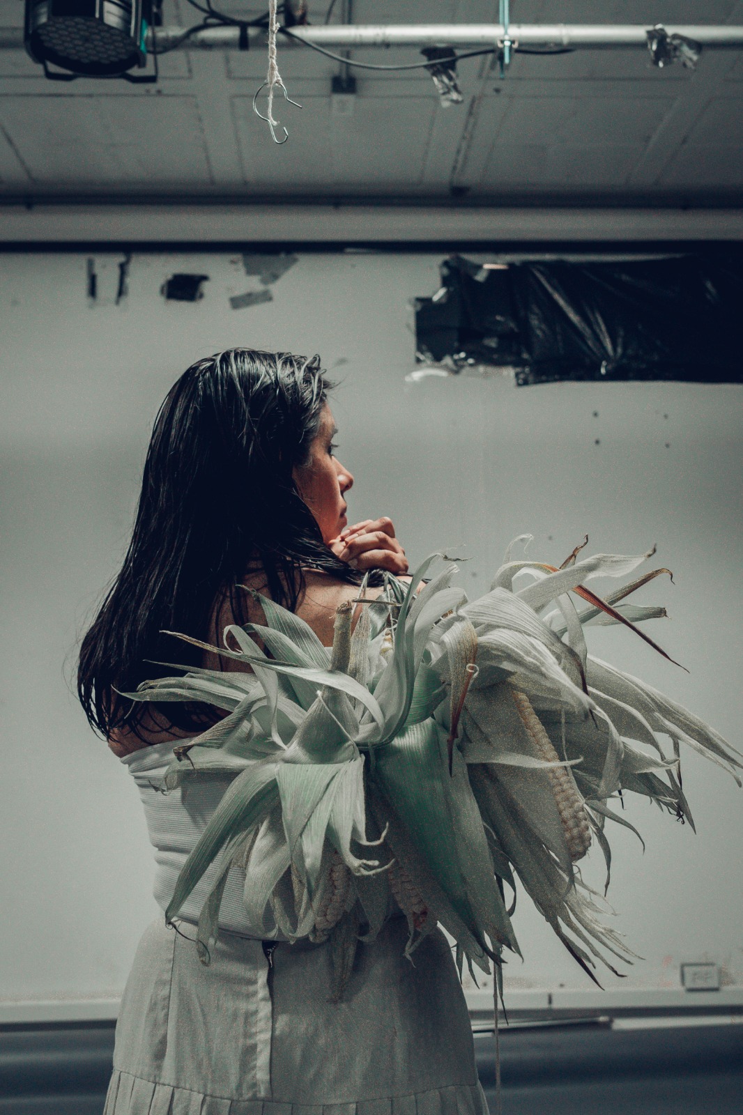 Foto de una joven de espaldas, ataviada con un vestido blanco. con el pelo negro suelto y que carga a su espalda un atado de varias mazorcas de maíz.