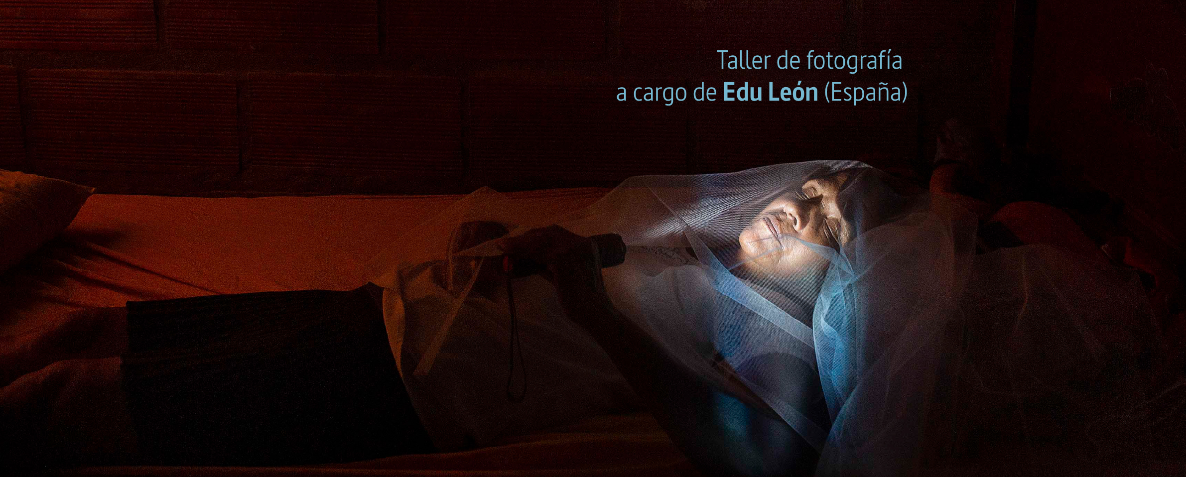Foto de una mujer acostada sobre una cama en un cuarto oscuro. Lleva sobre la cara un velo y se alumbra la cara con una linterna. En la parte superior de la foto se lee Taller de fotografía a cargo de Edu León (España).