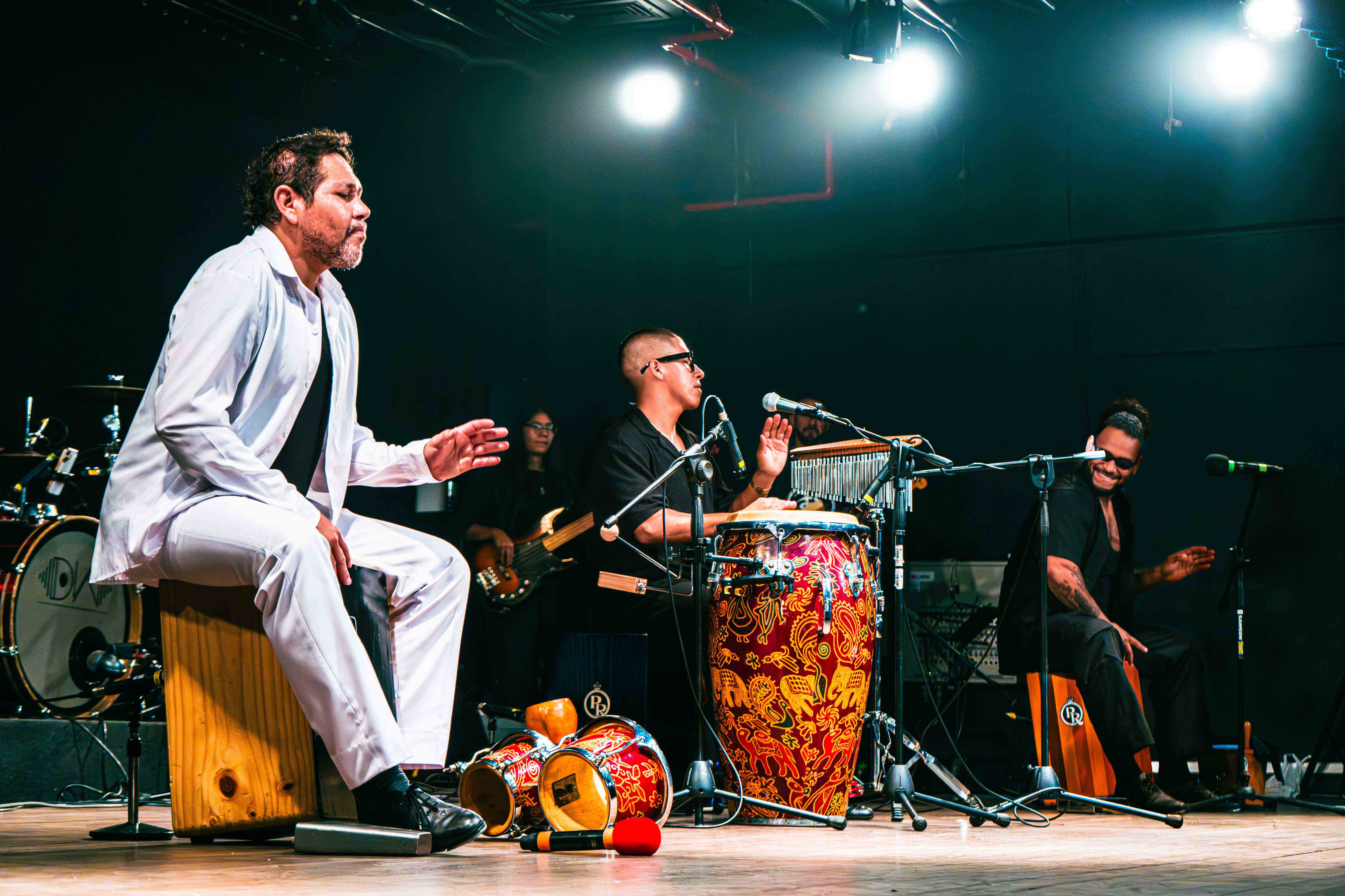 Foto que muestra en un escenario a tres músicos percusionistas: dos de ellos tocan cajón y el del medio toca una tumbadora. Los tres tienen micrófonos delante y otros instrumentos en el suelo, como bongoes. En el fondo se ve a una chica tocando guitarra.