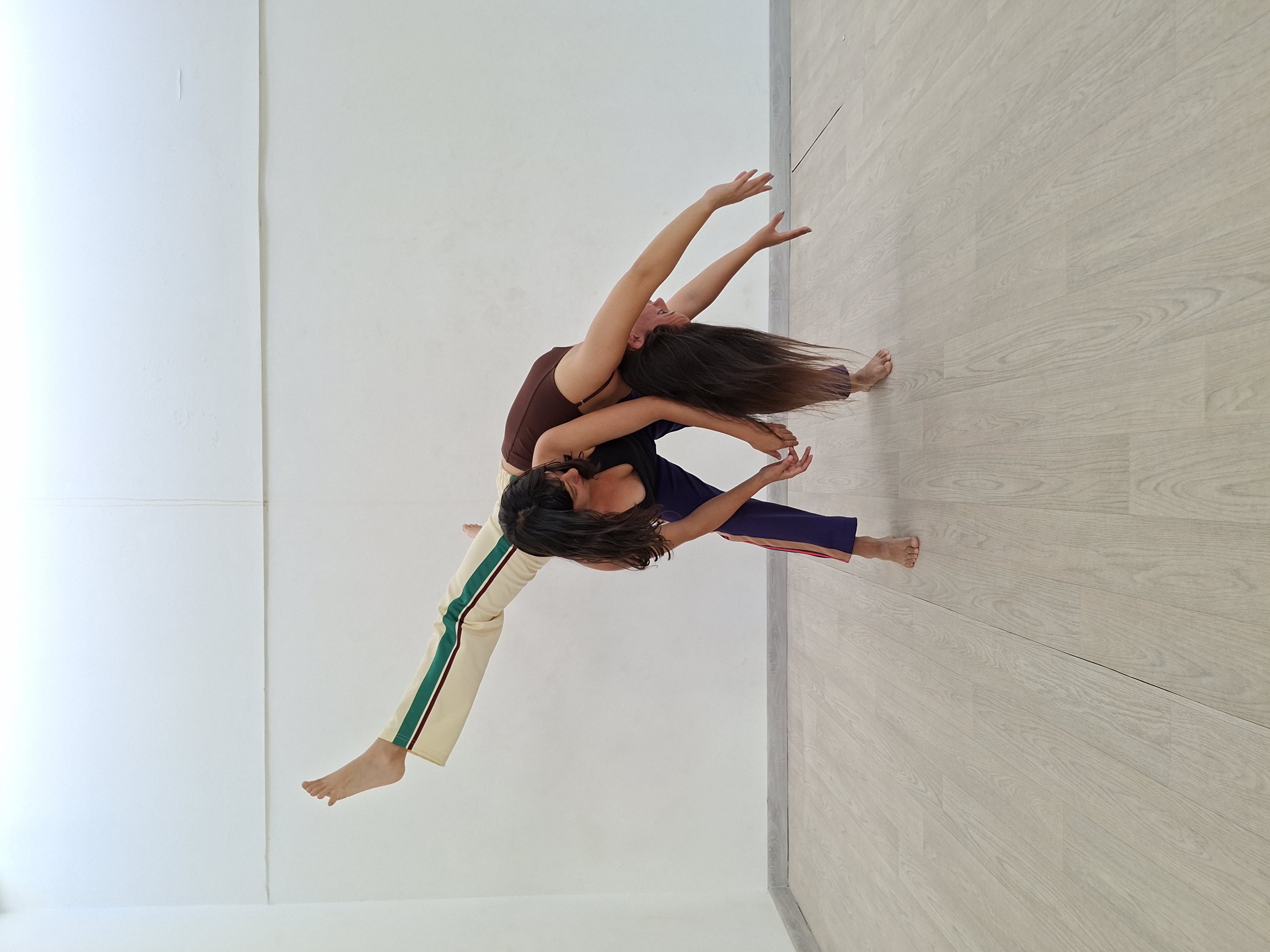 Foto que muestra a dos bailarinas en ropa de entrenamiento realizando un movimiento de danza en el que una sostiene sobre su espalda curvada hacia adelante el cuerpo de la otra.