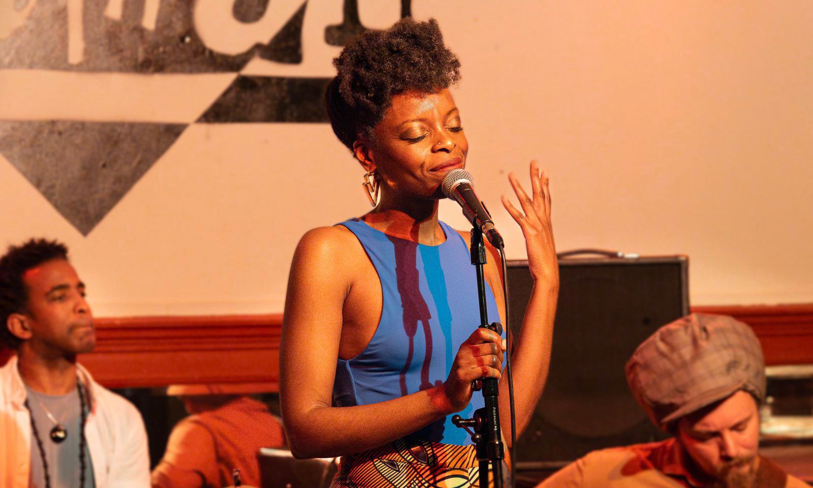 Foto de una cantante afrodescendiente interpretando alguna canción ante un micrófono y acompañada por un guitarrista y una percusionista.