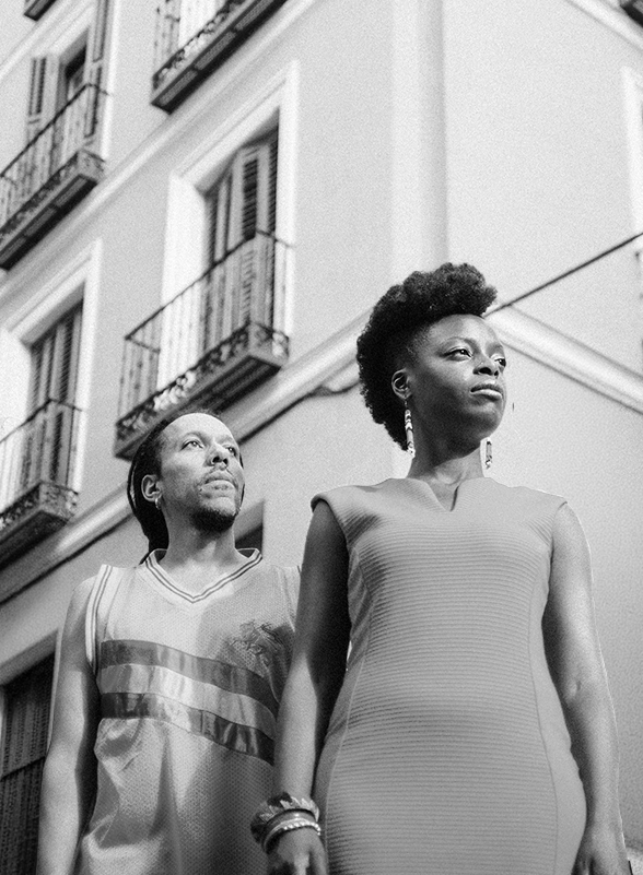 Foto en blanco y negro que muestra en primer plano a los artistas Astrid Jones y Carlos Bratt, parados en una calle mirando al horizonte. Detrás de ellos se divisan los balcones y puertas de la calle.