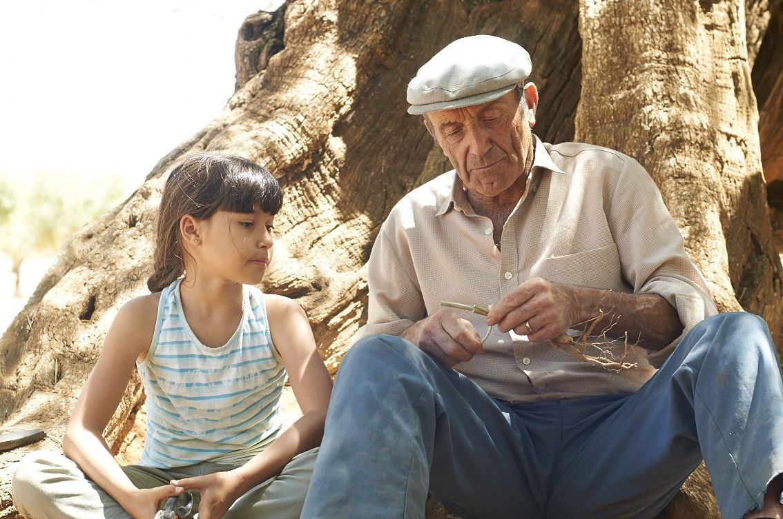 Foto de un señor mayor y una niña sentadoso en las raíces de un gran árbol. El señor sostiene una rama con la que le muestra algo a la niña.