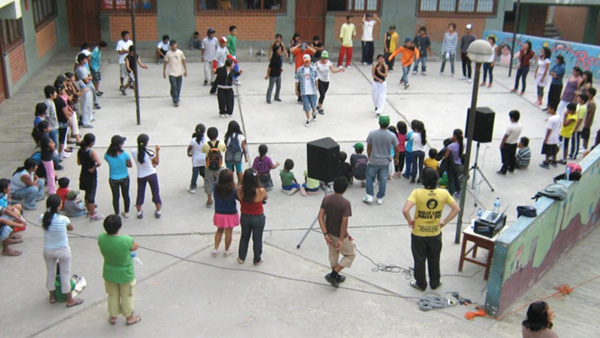 Foto que muestra el patio de un colegio en el que adultos y niños realizan actividades de juego y baile mientras otros los rodean observándoles.