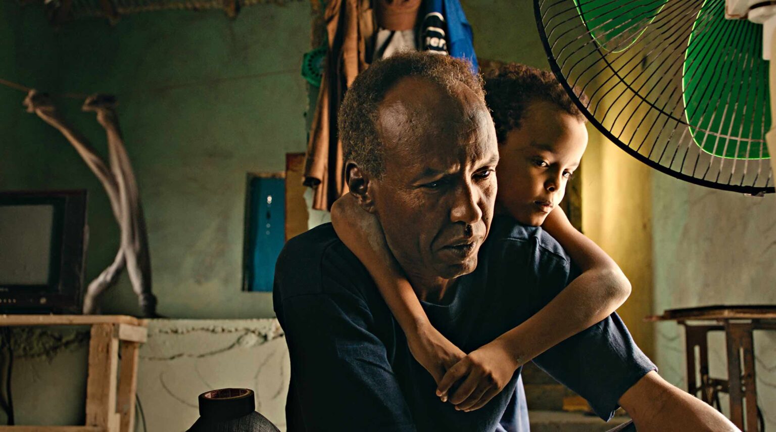Foto de un señor africano que caiga en intenta arreglar algo dentro su casa pobre, mientras su hijo, un niños de unos 10 años, se abrazo a él por la espalda