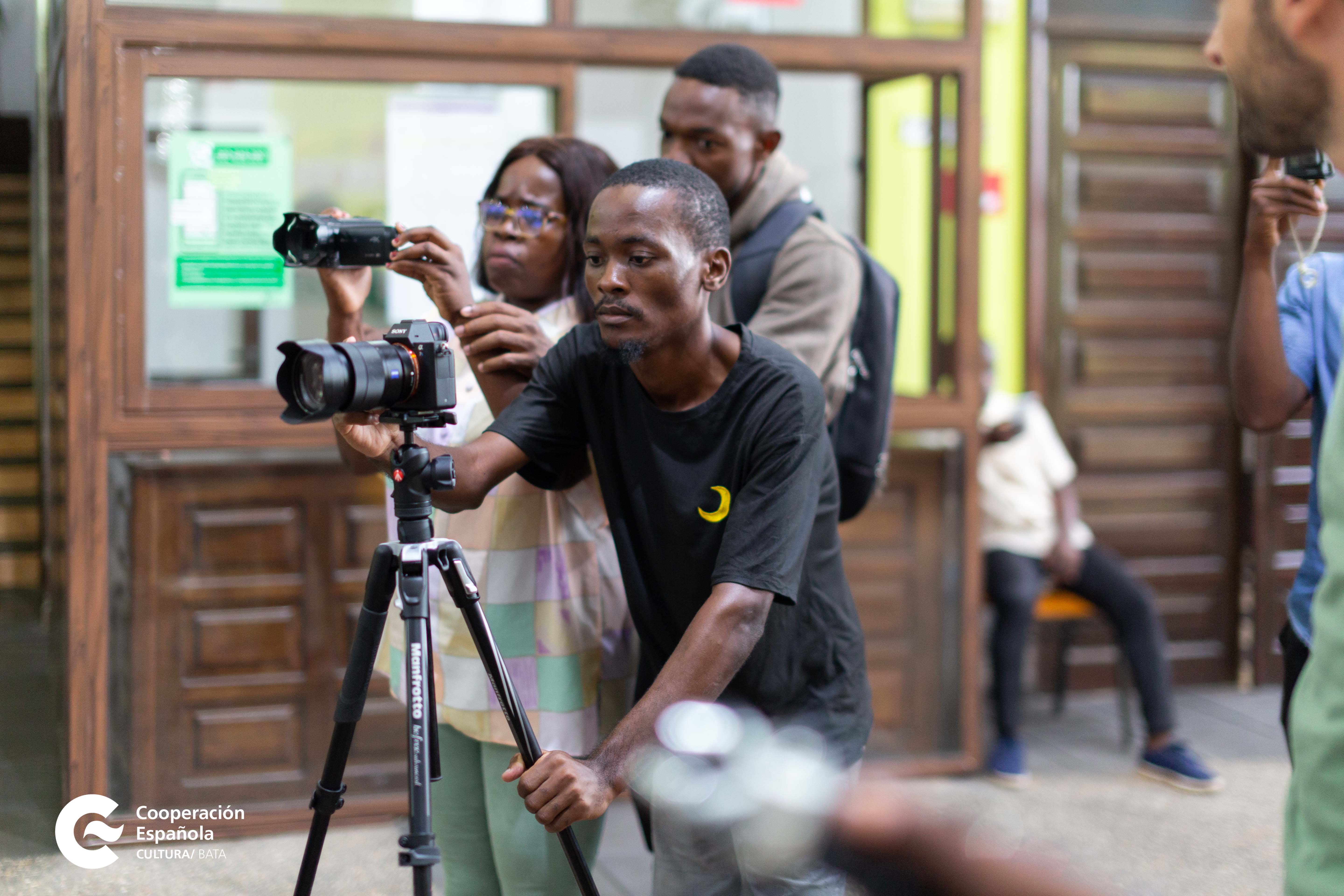 Foto que muestra en primer plano a un joven que está grabando alguna escena con una cámara apoyada sobre un trípode. En segundo plano, se ve a otra joven sosteniendo entre sus manos una cámara con la que graba y detrás de ella un joven también observa la escena que ella graba.